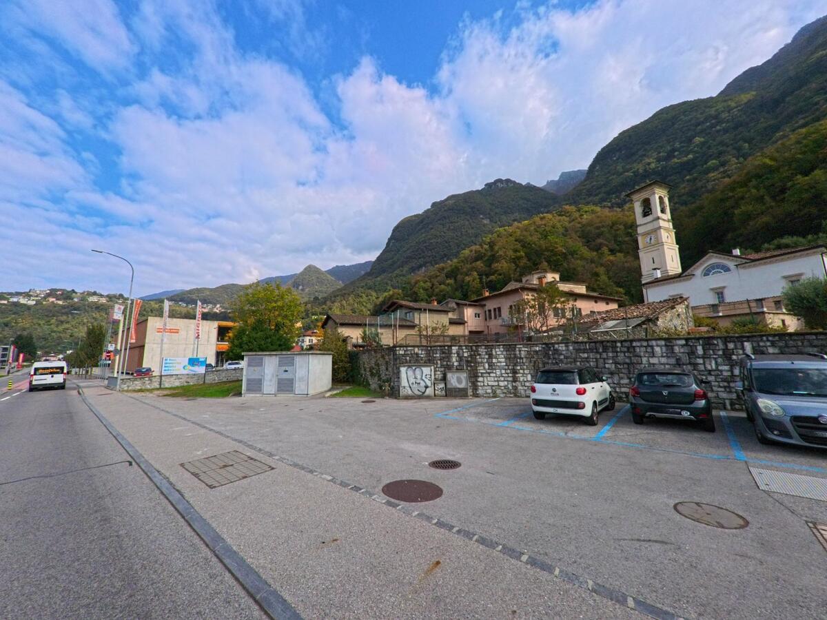 Ein Parkplatz mit abgestellten Autos liegt vor einer Steinmauer und Wohnhäusern in Melano (6818, Kanton Tessin), während der Kirchturm vor bewaldeten Berghängen aufragt.