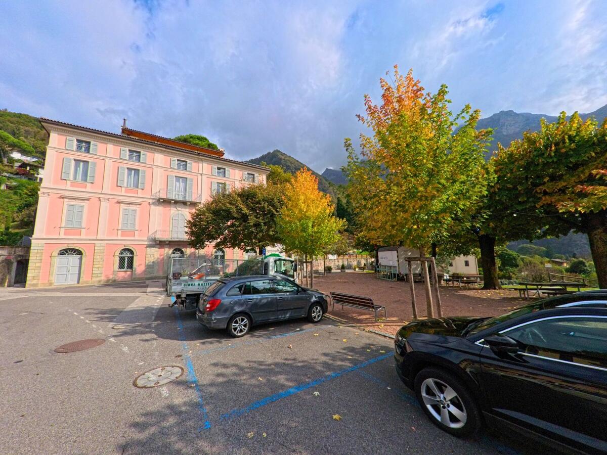 Ein Platz mit parkenden Autos und herbstlichen Bäumen liegt vor einem rosafarbenen Gebäude in Melano (6818, Kanton Tessin) vor einer bewaldeten Bergkulisse.