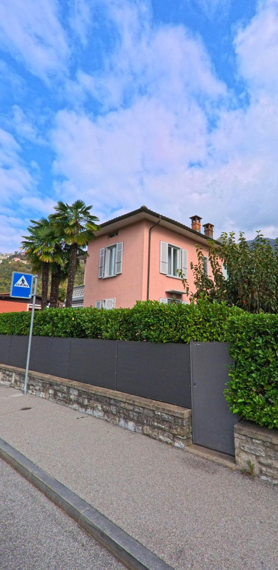 Hinter einer Hecke und einer niedrigen Mauer steht ein rosafarbenes Wohnhaus mit Palmen in Melano (6818, Kanton Tessin) an einer ruhigen Strasse.