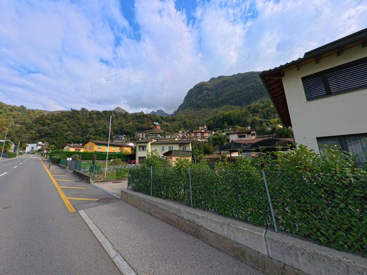 Eine Strasse mit Gehweg führt durch ein Wohnquartier in Melano (6818, Kanton Tessin), während sich dahinter dicht bewaldete Hügel mit verstreuten Häusern erheben.