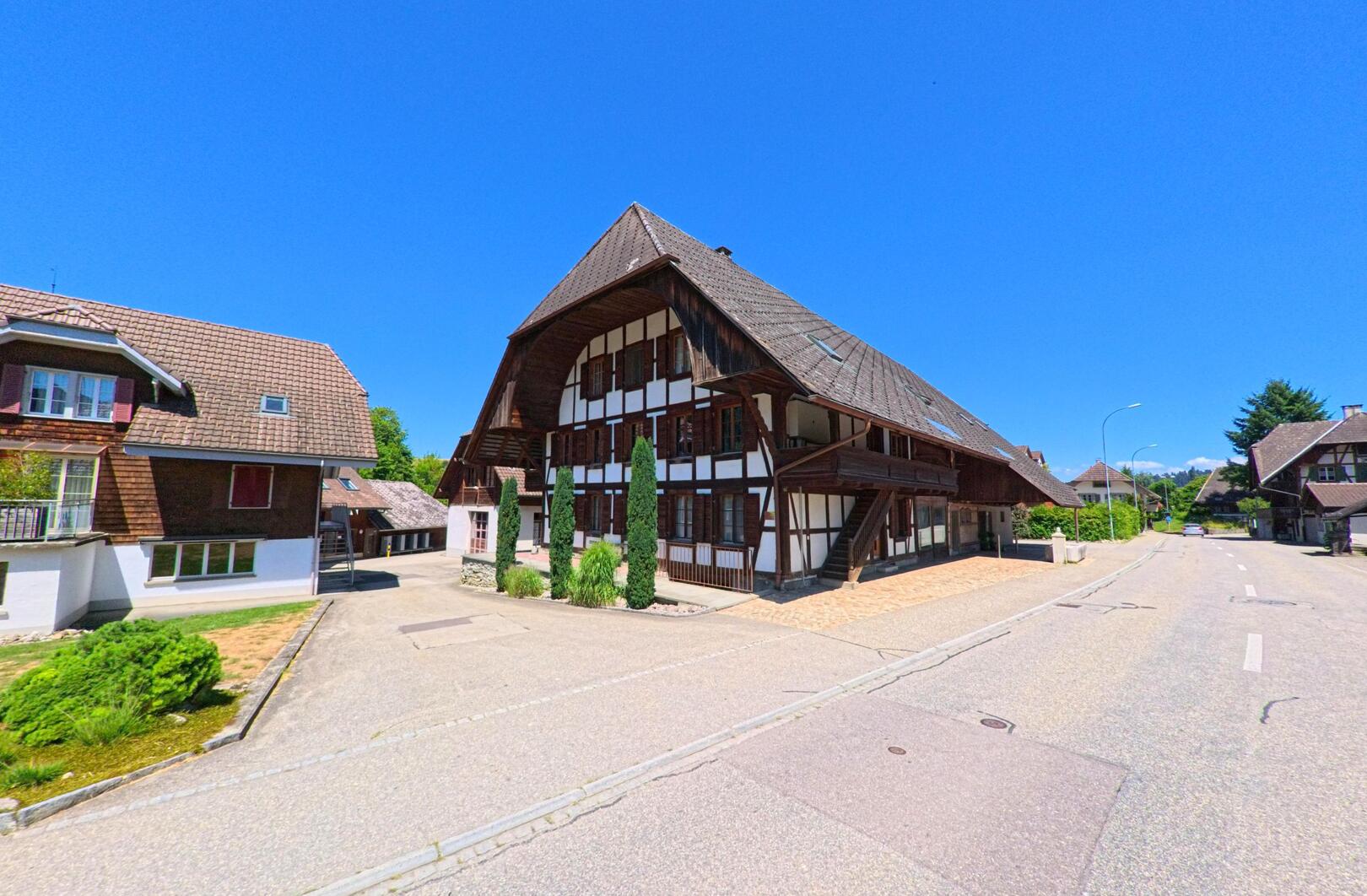 Ein imposantes Bauernhaus mit markanter Fachwerkfassade praegt das Ortsbild von Melchnau (4917, Kanton Bern) entlang einer ruhigen Strasse mit weiteren Wohngebaeuden.