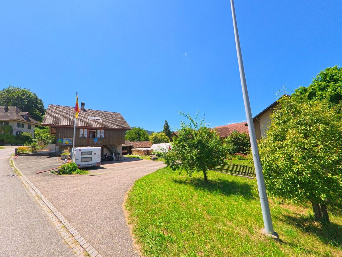 Ein Holzhaus mit Schweizer Fahne und geparktem Wohnwagen steht in Melchnau (4917, Kanton Bern) an einer ruhigen Seitenstrasse mit angrenzender Gruenflaeche und Obstbaeumen.