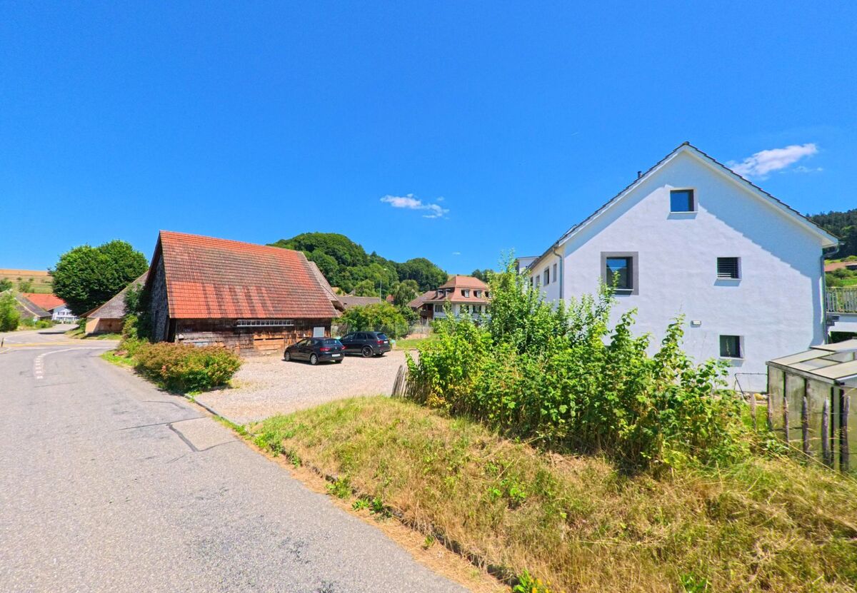 In Melchnau (4917, Kanton Bern) trifft ein moderner weisser Wohnbau mit Garten auf eine traditionelle Scheune mit Ziegeldach entlang einer Landstrasse am Siedlungsrand.