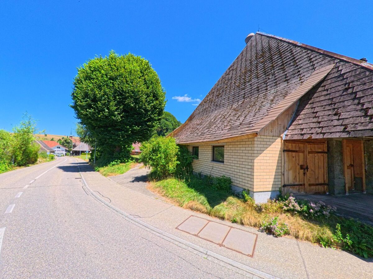 Ein Holzgebaeude mit steilem Walmdach steht in Melchnau (4917, Kanton Bern) an einer sanft ansteigenden Landstrasse, gesaeumt von Baeumen und Gruenflaechen.