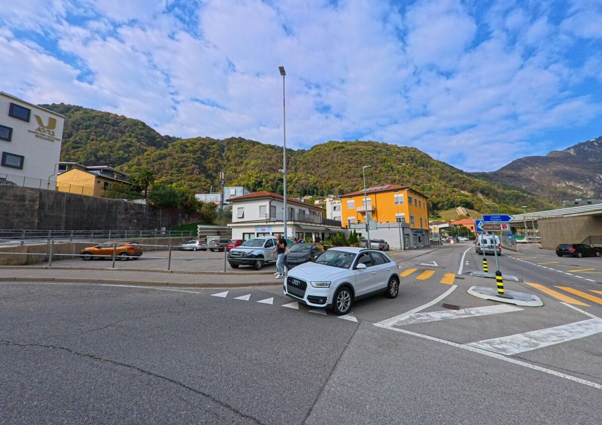 An einer belebten Kreuzung in Melide (6815, Kanton Tessin) fahren Autos vor dem Hintergrund bewaldeter Huegel und farbiger Gebaeude entlang.