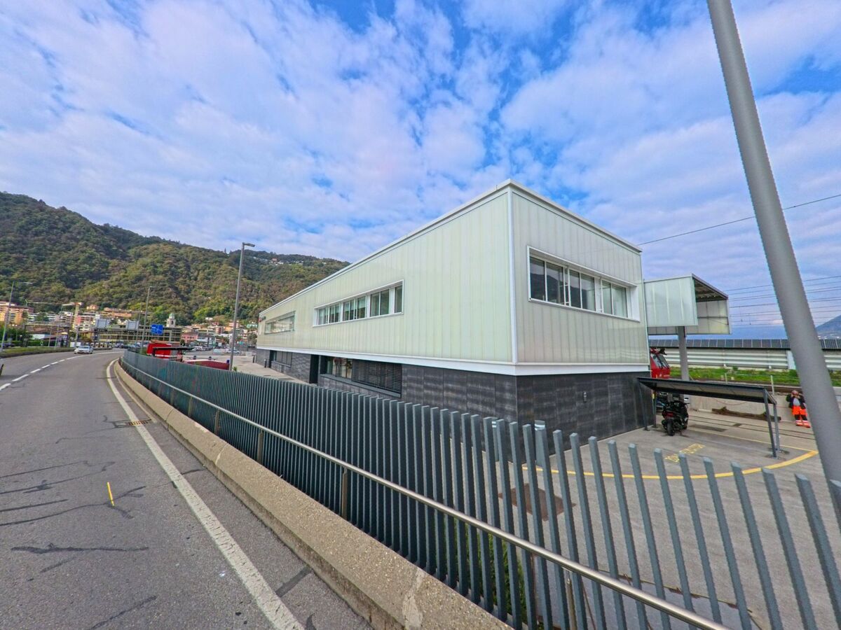 Ein modernes, helles Gebaeude steht in Melide (6815, Kanton Tessin) neben den Bahngleisen, mit Blick auf die Strasse und die umliegenden Huegel.