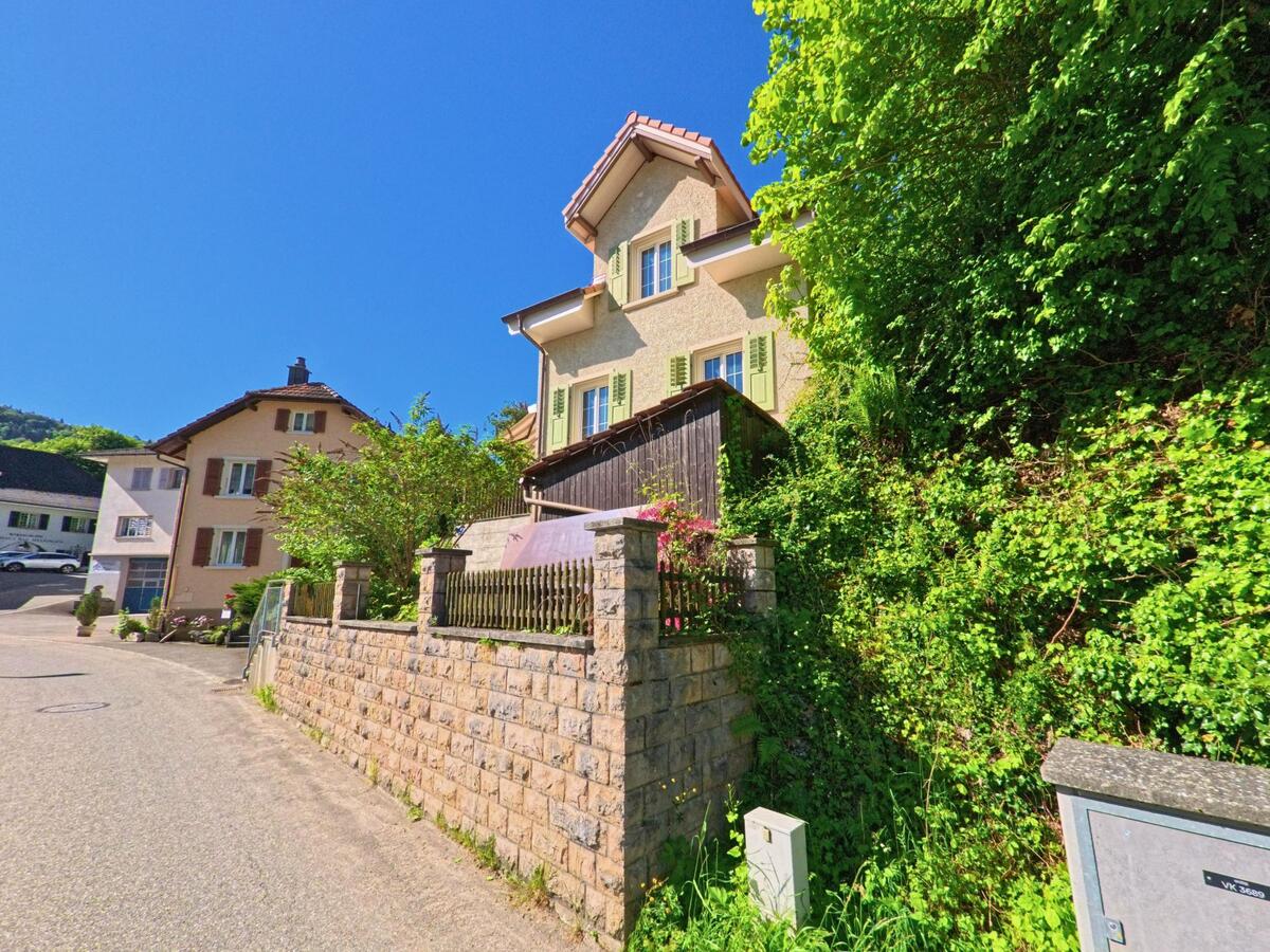 In Meltingen (4233, Kanton Solothurn) säumen gepflegte Einfamilienhäuser mit kleinen Vorgärten eine ruhige, leicht ansteigende Quartierstrasse am Waldrand.