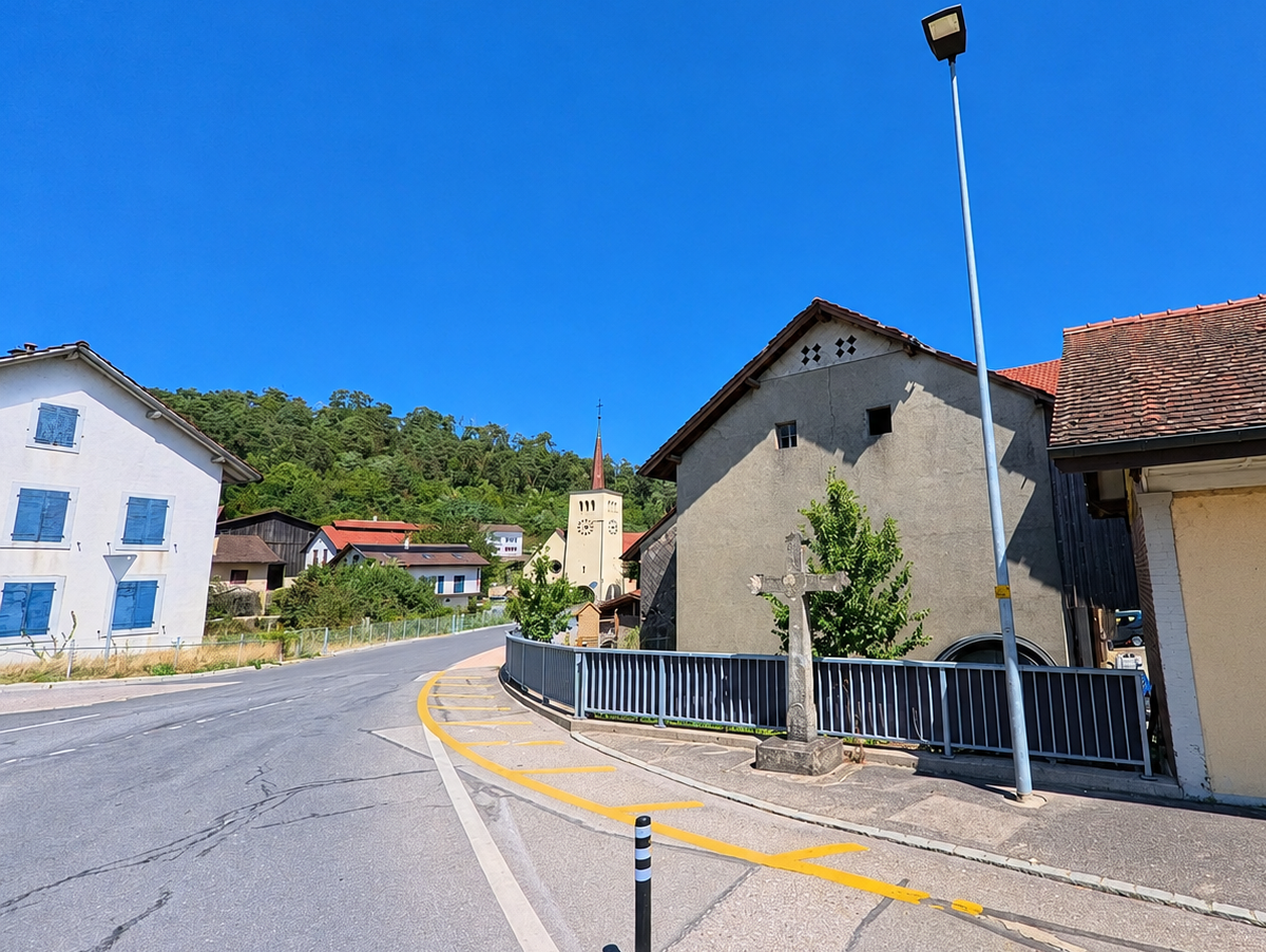 Eine Strasse in Ménières (1533, Kanton Freiburg) führt an mehreren traditionellen Gebäuden vorbei in Richtung eines Kirchturms, während ein steinernes Kreuz am Strassenrand steht.