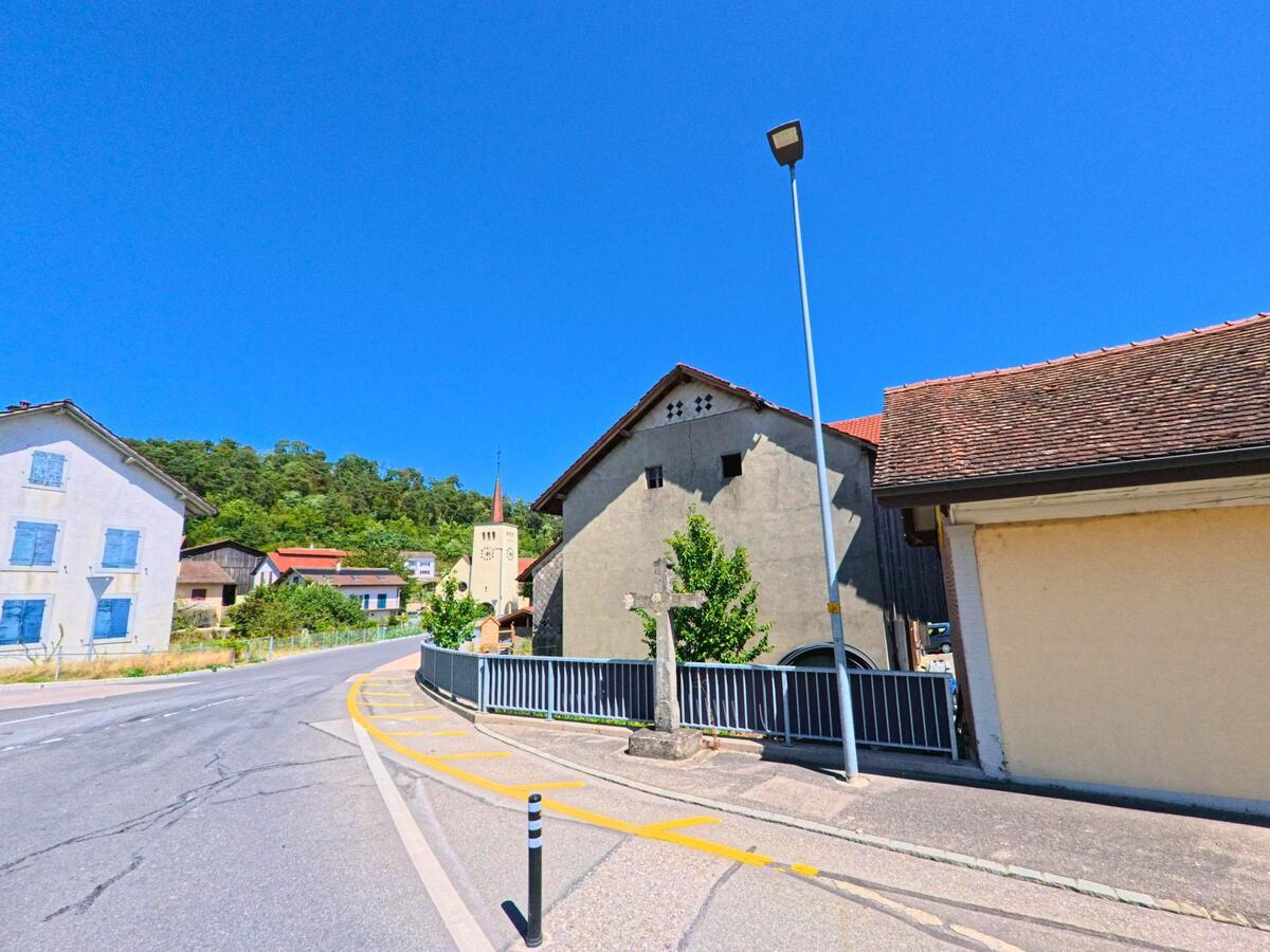 In Ménières (1533, Kanton Freiburg) verläuft eine Strasse an alten Gebäuden vorbei, im Hintergrund ragt der Kirchturm mit spitzem Dach auf.