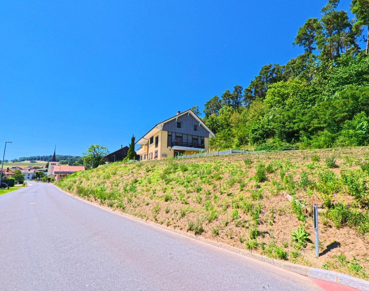 An einer leicht ansteigenden Strasse in Ménières (1533, Kanton Freiburg) steht ein gelbes Wohnhaus am Waldrand, im Hintergrund ist der Kirchturm zu sehen.
