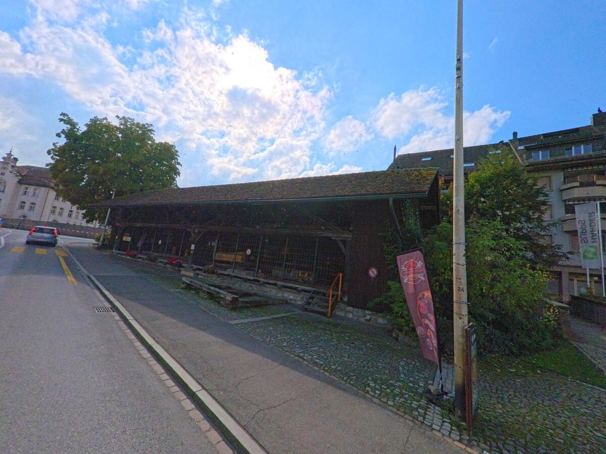 Entlang der Hauptstrasse in Menziken (5736, Kanton Aargau) steht ein langes Holzgebäude mit traditionellem Dach, neben dem ein Auto in Richtung Zentrum fährt.