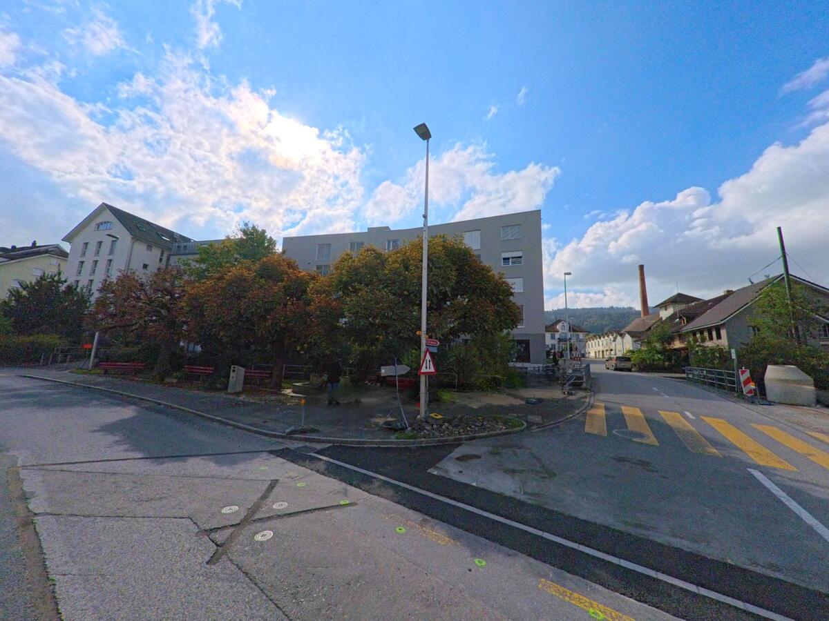 An einer Strassenkreuzung in Menziken (5736, Kanton Aargau) stehen moderne und ältere Gebäude nebeneinander, während herbstlich gefärbte Bäume den Platz säumen.