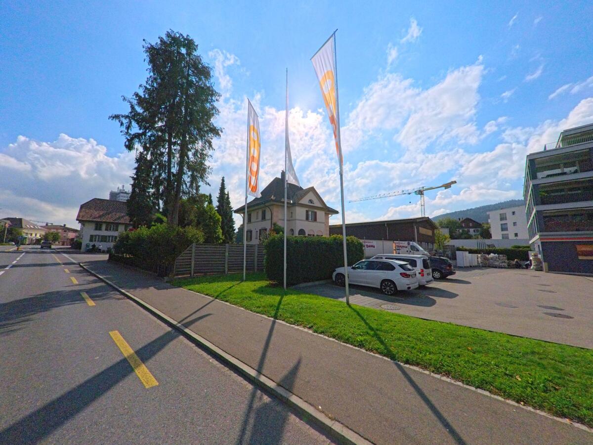 An einer Strasse in Menziken (5737, Kanton Aargau) wehen hohe Fahnen neben einem Parkplatz und modernen Gebäuden, im Hintergrund ist ein Baukran zu sehen.