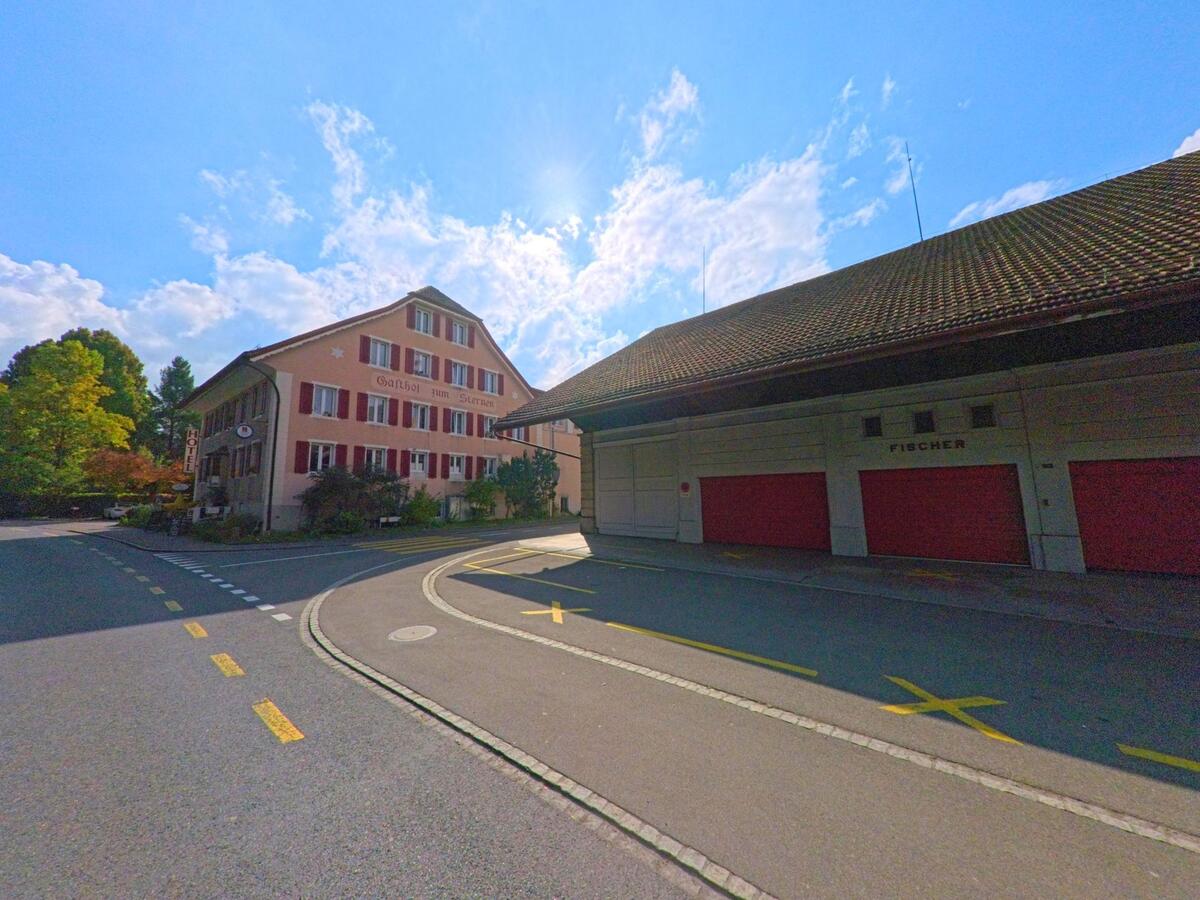 An einer Kurve in Menziken (5737, Kanton Aargau) steht der Gasthof zum Sternen mit roten Fensterläden und einem grossen Schriftzug an der Fassade.