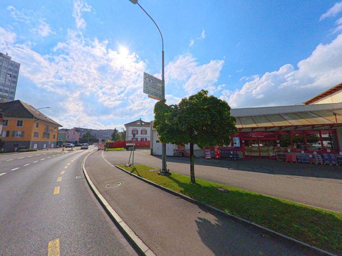 An einer sonnigen Hauptstrasse in Menziken (5737, Kanton Aargau) liegt ein rotes Geschäft mit Glasfassade, während Autos in Richtung Ortszentrum fahren.