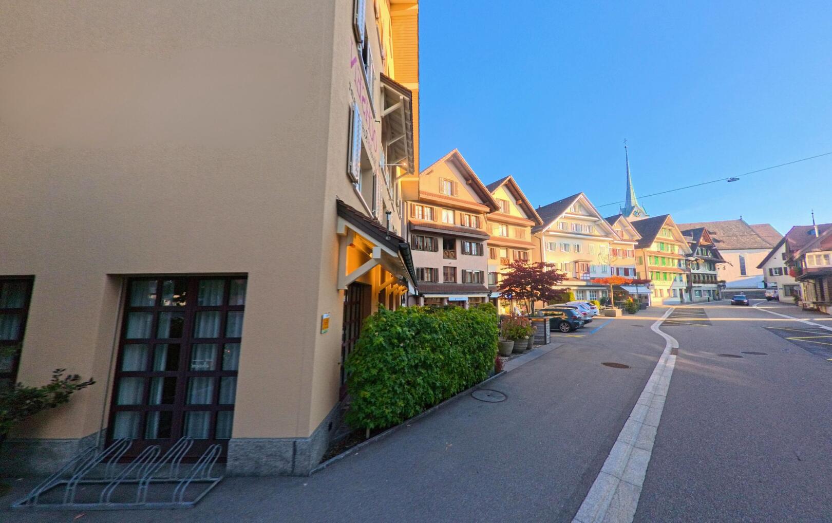 In der Ortsmitte von Menzingen (6313, Kanton Zug) zeigt das Bild eine sonnige Strasse mit traditionellen Hausfassaden und einem Kirchturm im Hintergrund.