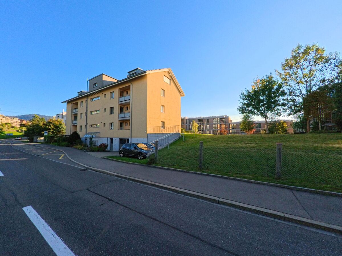 In Menzingen (6313, Kanton Zug) steht ein helles Mehrfamilienhaus an einer leicht ansteigenden Strasse, neben einer offenen Gruenflaeche mit einzelnen Baeumen.