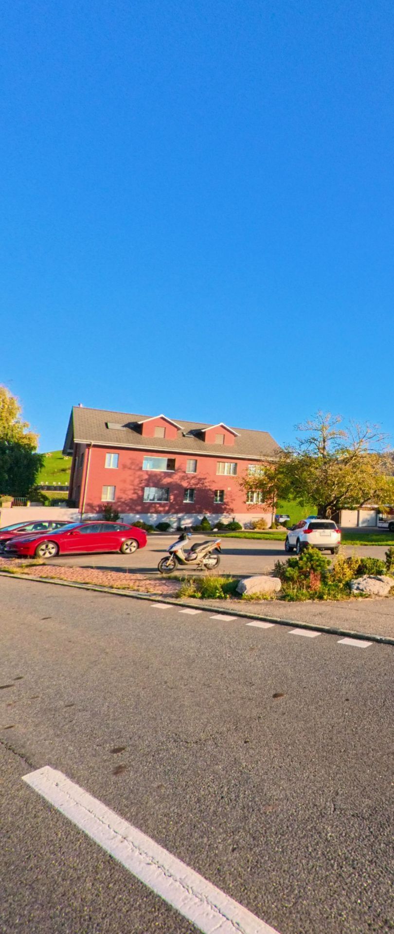 In Menzingen (6313, Kanton Zug) steht ein rotes Wohnhaus vor einer offenen Parkflaeche, auf der mehrere Autos und ein Motorroller im Sonnenlicht zu sehen sind.