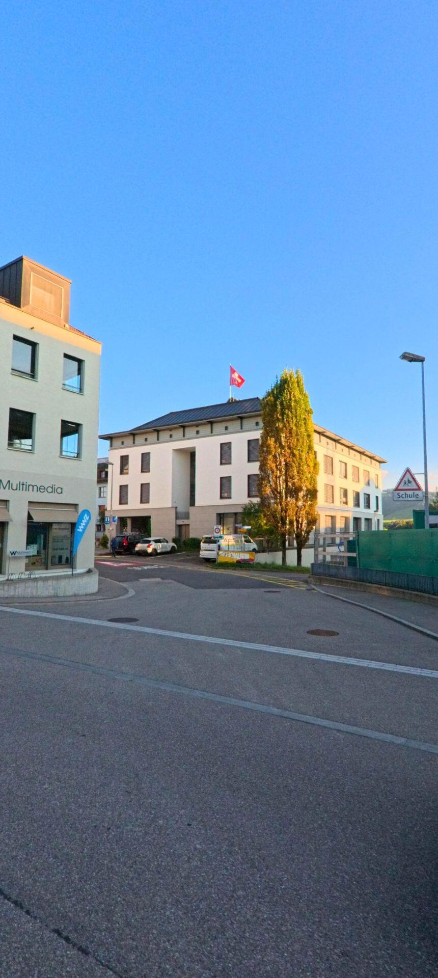 In Menzingen (6313, Kanton Zug) steht ein helles Verwaltungsgebäude mit Schweizer Flagge an einer Einmündung, flankiert von Baeumen und umliegenden Gewerbebauten.