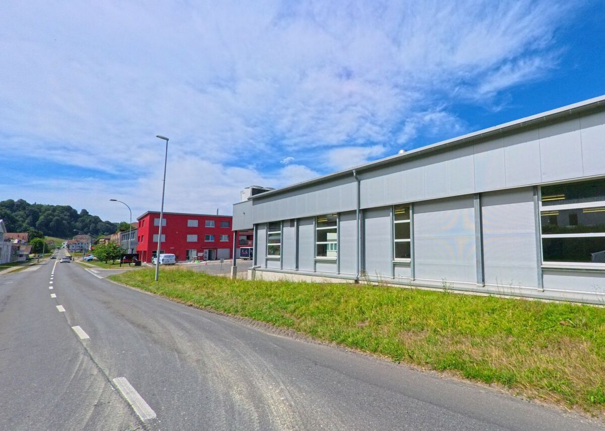 In Menznau (6122, Kanton Luzern) verläuft eine Strasse an einem flachen Industriegebaeude mit grossen Fenstern vorbei, im Hintergrund hebt sich ein rotes Verwaltungsgebaeude deutlich ab.