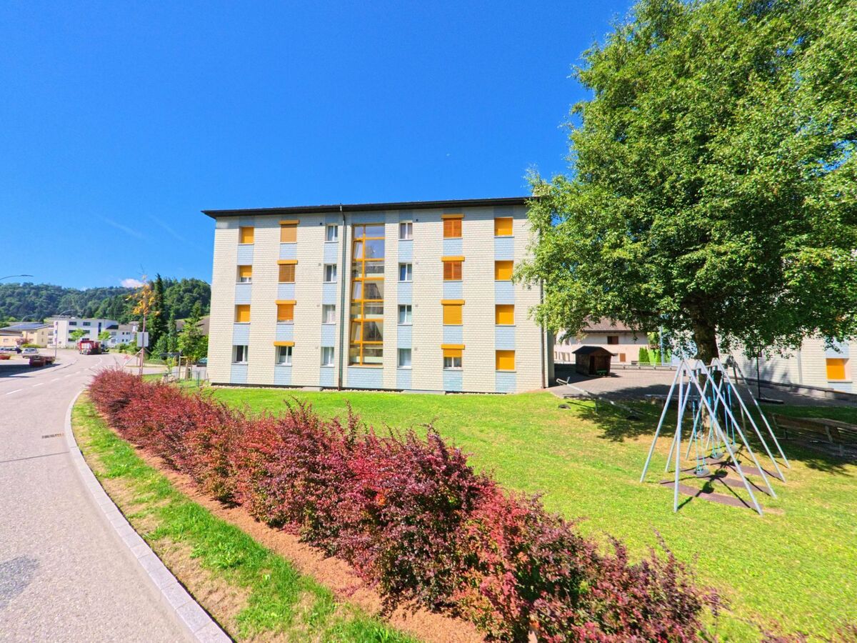  In Menznau (6122, Kanton Luzern) steht ein mehrstöckiges Wohnhaus mit gelben Fensterlaeden an einer ruhigen Strasse, daneben befindet sich eine kleine Spielplatzanlage unter einem grossen Baum.