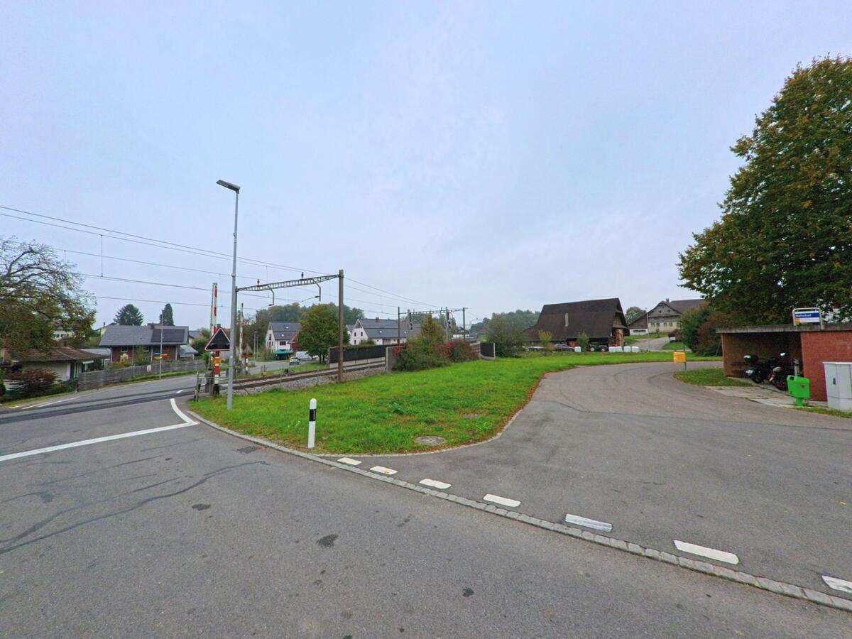 Eine Bahnlinie mit Schranken kreuzt eine Strasse in Merenschwand (5634, Kanton Aargau), im Hintergrund sind Wohnhäuser und landwirtschaftliche Gebäude zu sehen.