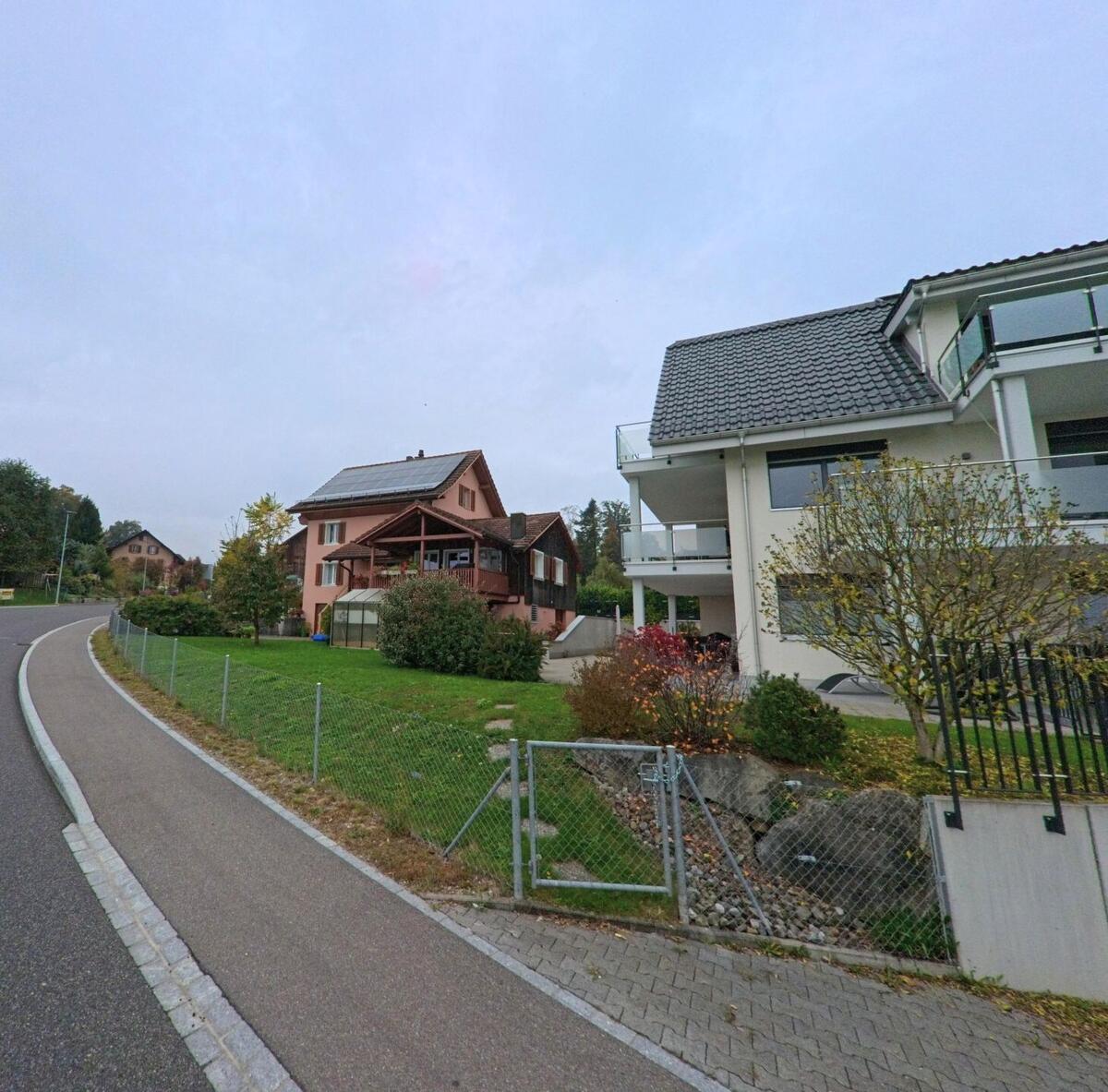 In einem Wohngebiet von Merenschwand (5634, Kanton Aargau) stehen Einfamilienhäuser mit gepflegten Gärten an einer leicht ansteigenden Strasse unter grauem Himmel.