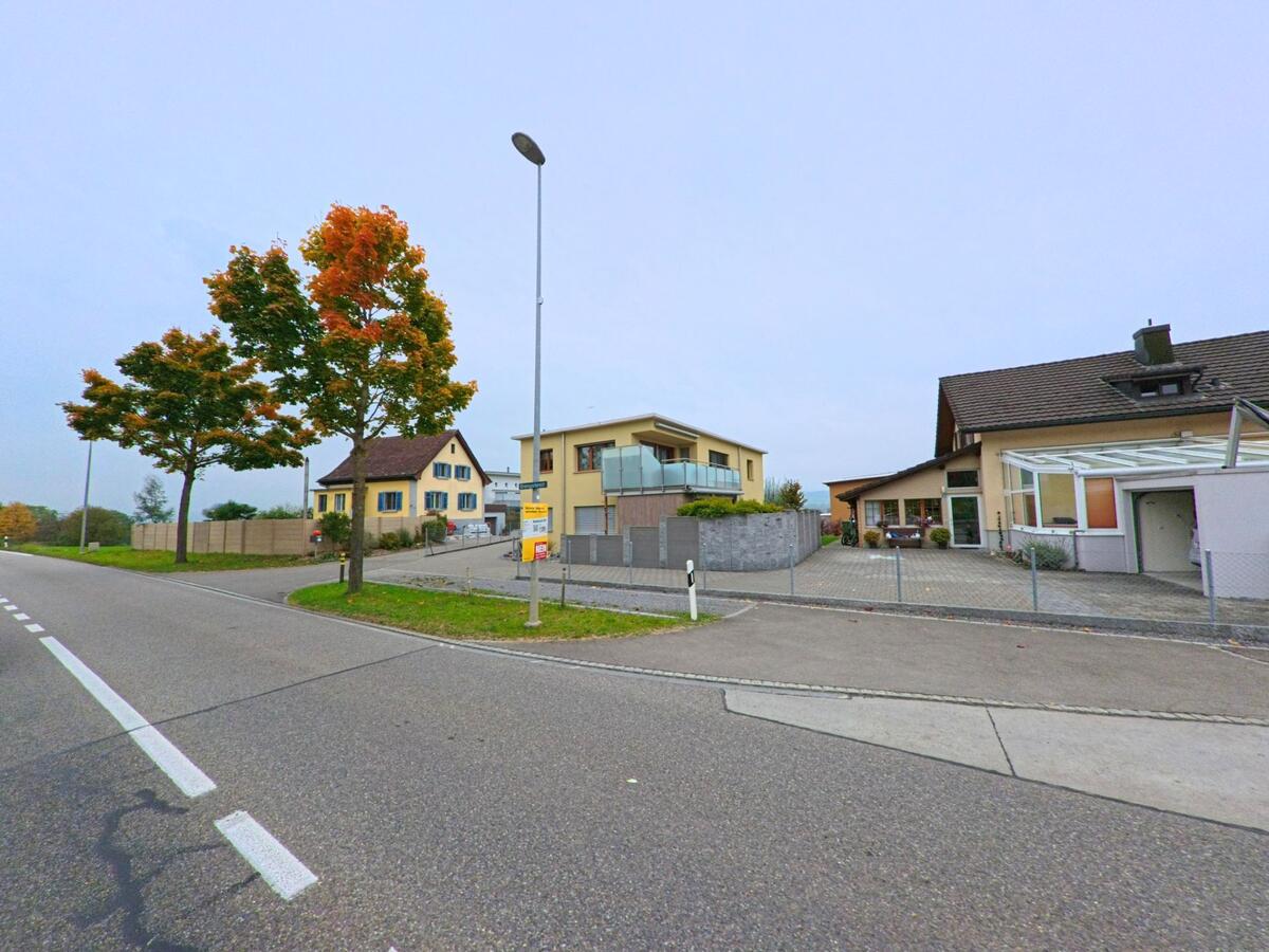 Entlang der Hauptstrasse in Merenschwand (5634, Kanton Aargau) stehen Einfamilienhäuser mit modernen und traditionellen Fassaden, flankiert von Bäumen mit herbstlich gefärbtem Laub.