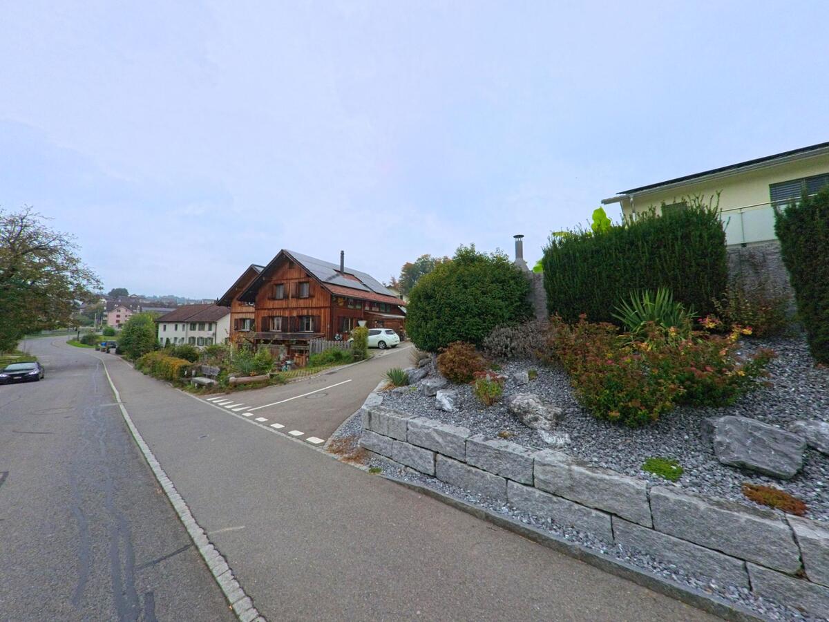An einer leicht ansteigenden Strasse in Merenschwand (5634, Kanton Aargau) steht ein traditionelles Holzhaus mit gepflegtem Garten und moderner Böschungsgestaltung im Vordergrund.