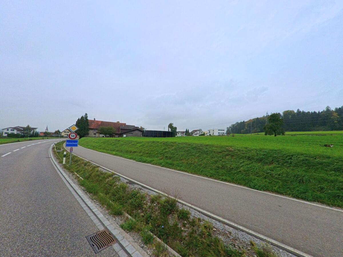 Am Ortseingang von Merenschwand (5634, Kanton Aargau) führt eine Landstrasse zwischen grünen Wiesen hindurch, begleitet vom Ortsschild und Bauernhäusern im Hintergrund.
