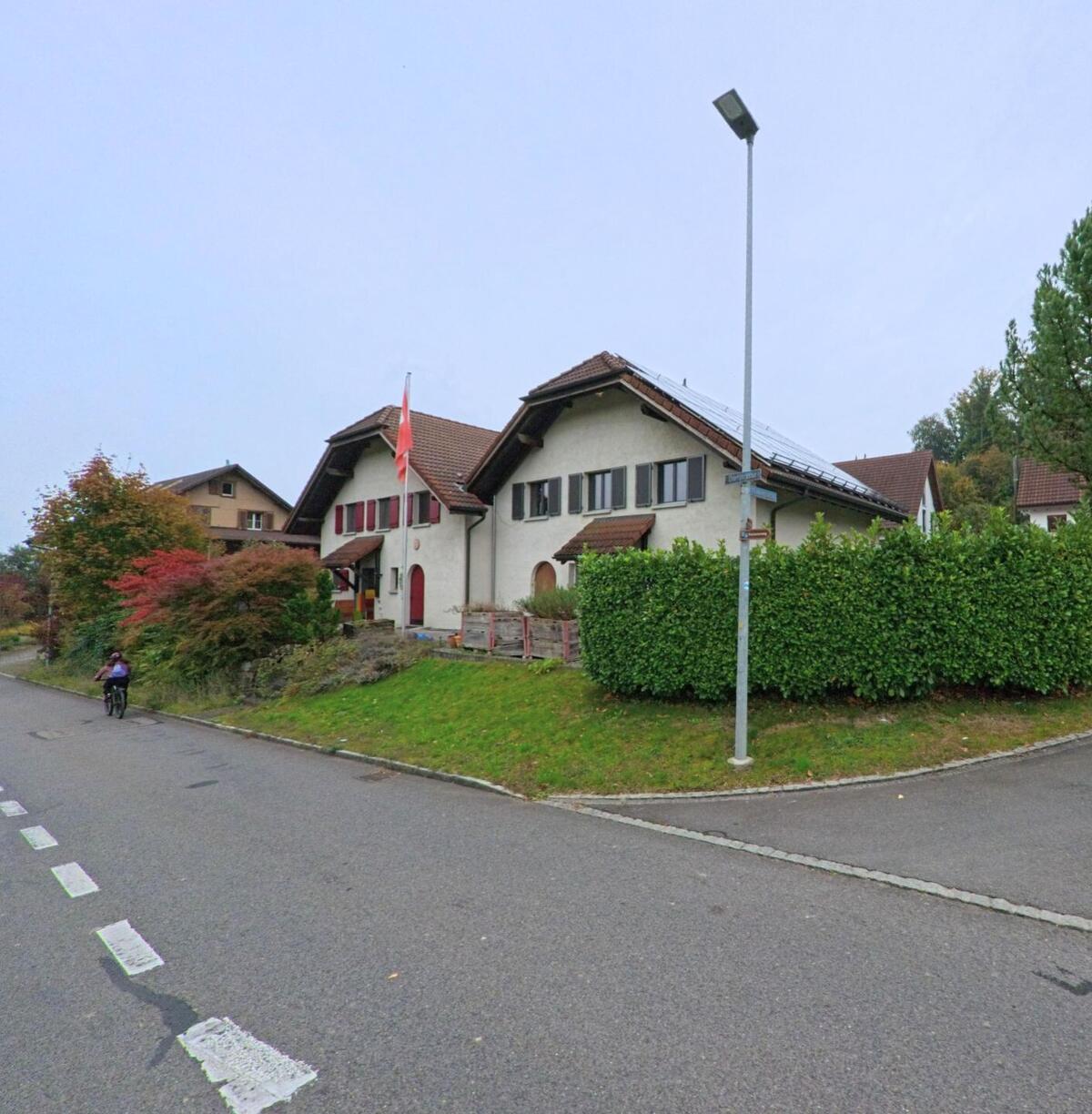 Ein Wohnhaus mit rotem Ziegeldach und Schweizer Fahne steht an einer Nebenstrasse in Merenschwand (5634, Kanton Aargau), daneben fährt eine Person mit dem Fahrrad vorbei.