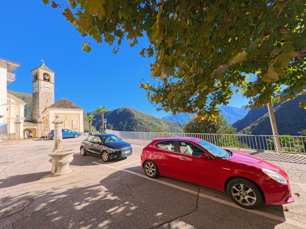 Auf dem Dorfplatz von Mergoscia (6647, Kanton Tessin) stehen Autos im Schatten eines Baumes, im Hintergrund die Kirche mit Glockenturm.