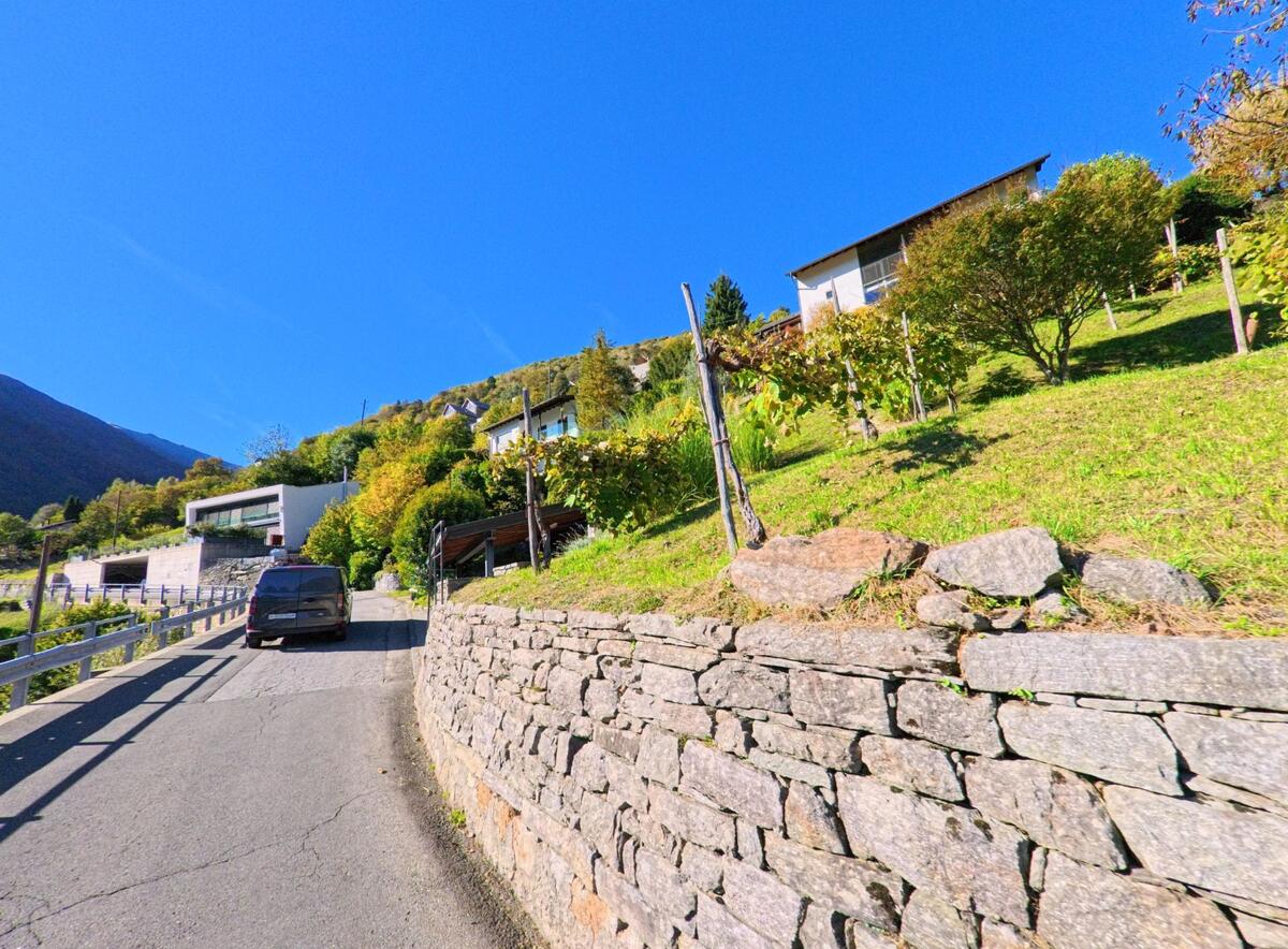 Eine schmale, ansteigende Strasse in Mergoscia (6647, Kanton Tessin) führt vorbei an modernen und traditionellen Häusern mit Gärten.