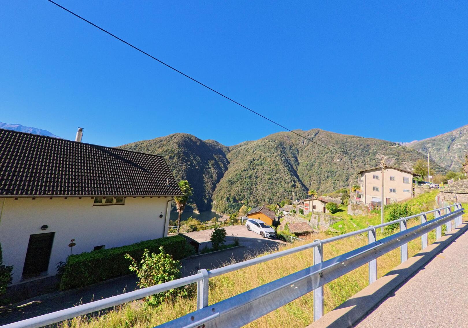 Entlang einer steilen Strasse in Mergoscia (6647, Kanton Tessin) stehen Häuser mit Blick auf bewaldete Hügel und den See.