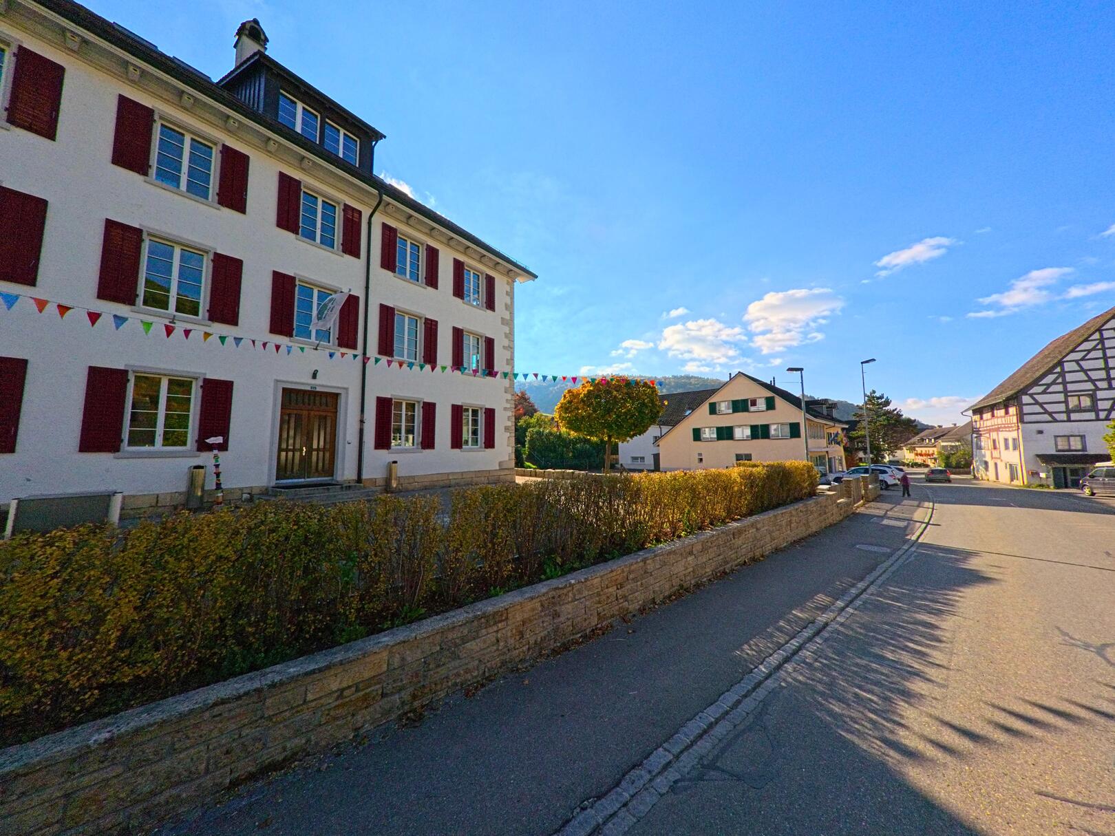 In Merishausen (8232, Kanton Schaffhausen führt eine Strasse an traditionellen Gebäuden entlang, die mit bunten Fahnen geschmückt sind, unter strahlend blauem Himmel.
