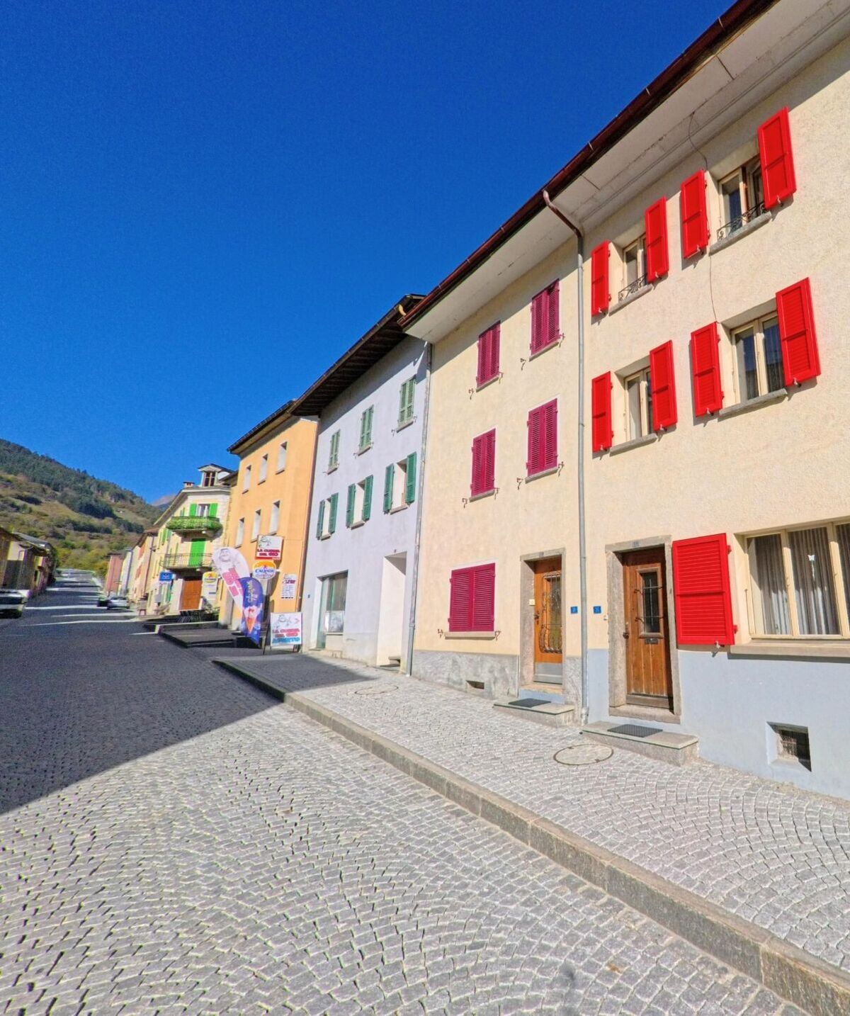 Entlang einer gepflasterten Dorfstrasse in Mesocco (6563, Kanton Graubünden) stehen farbig gestrichene Wohnhäuser mit Fensterläden unter einem klaren, tiefblauen Himmel.