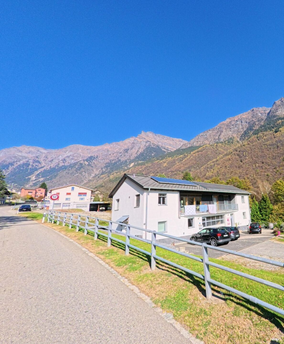 An einer ruhigen Strasse in Mesocco (6563, Kanton Graubünden) steht ein modernes Wohnhaus mit Balkon, während sich dahinter bewaldete Hänge und felsige Berge unter klarem Himmel erheben.