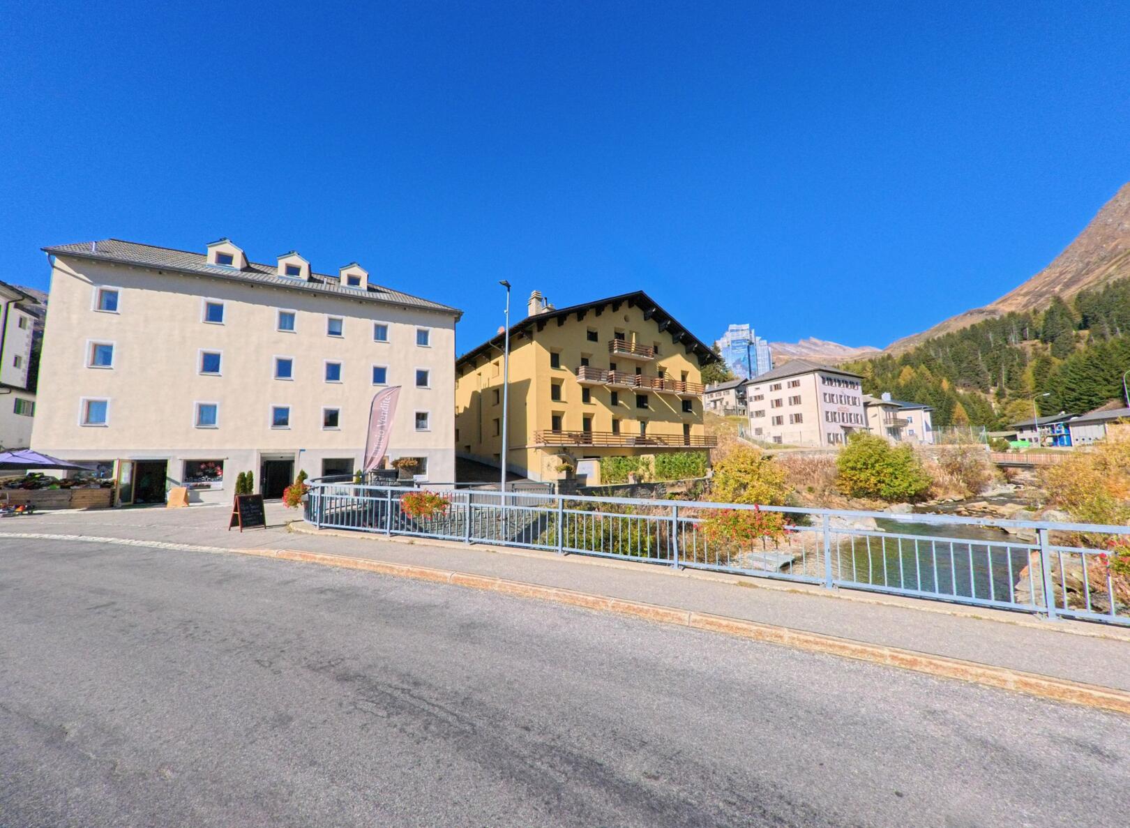 Über eine Brücke hinweg zeigt sich in Mesocco (6565, Kanton Graubünden) eine Reihe von Wohn- und Hotelgebäuden entlang eines Bachlaufs vor sonniger Bergkulisse.