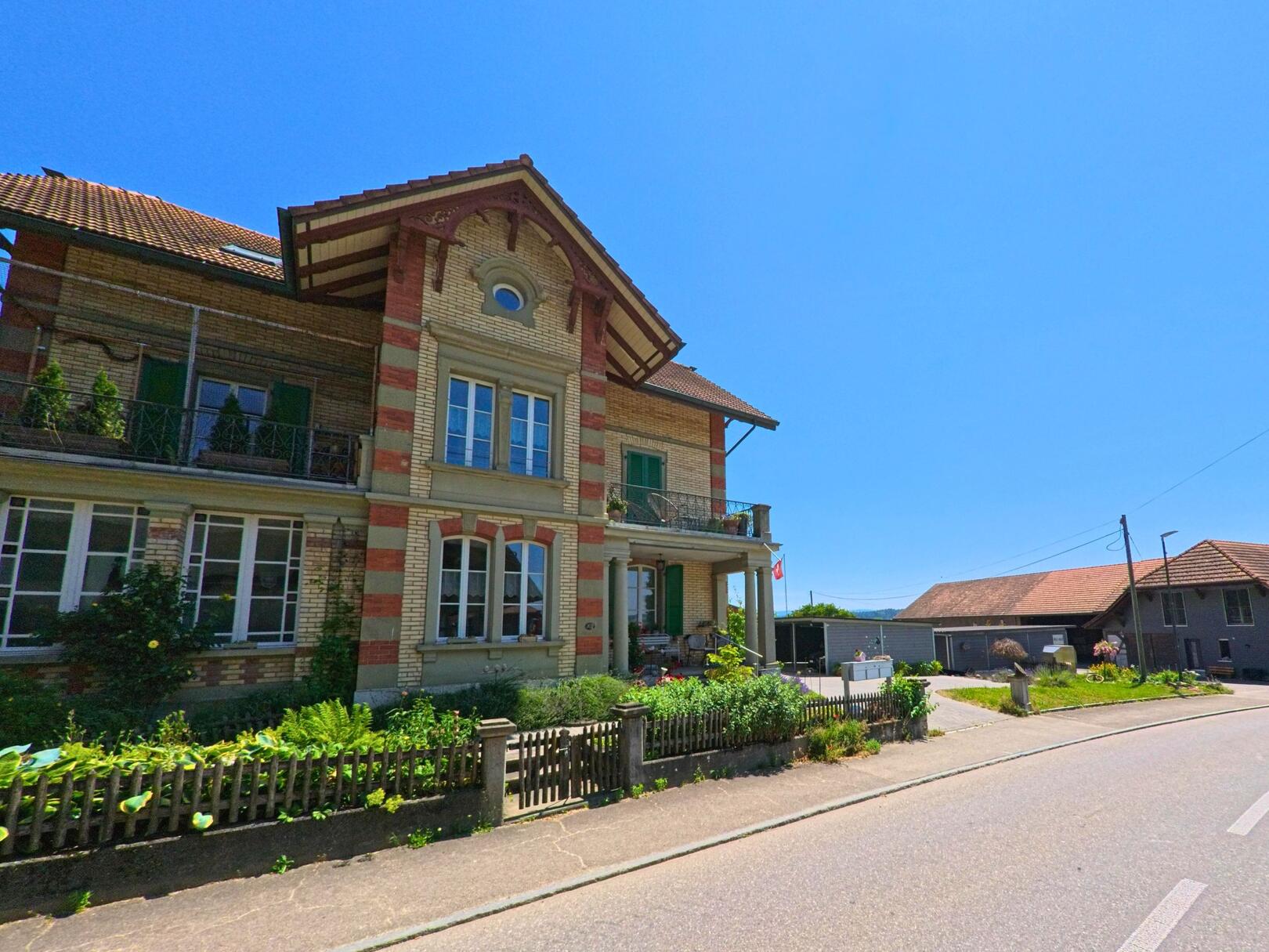In Messen (3254, Kanton Solothurn) steht ein historisches Wohnhaus aus Backstein mit Balkonen und Vorgarten direkt an einer wenig befahrenen Strasse.