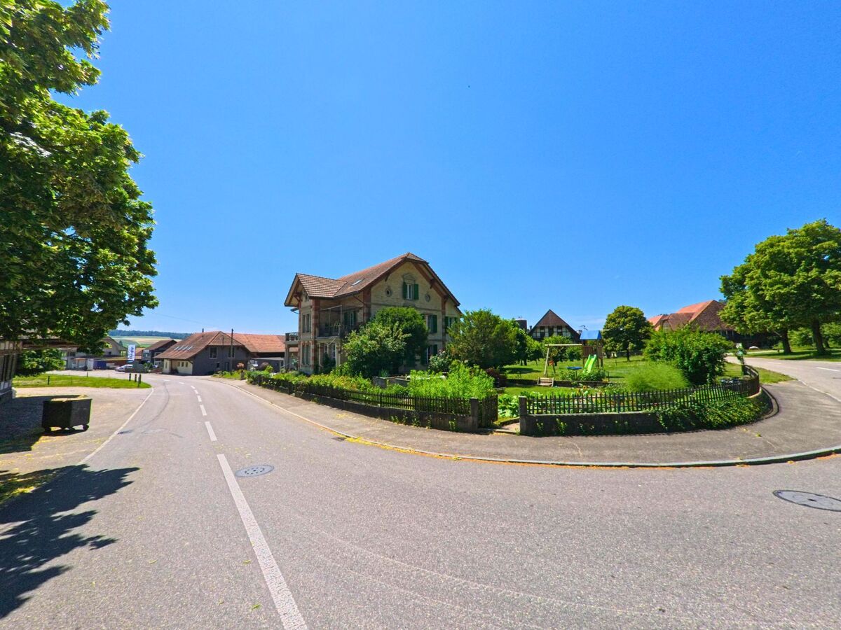 In Messen (3254, Kanton Solothurn) liegt ein altes Wohnhaus mit Gartenanlage an einer sanften Kurve im Dorf, umgeben von weiteren Bauernhäusern und Bäumen.