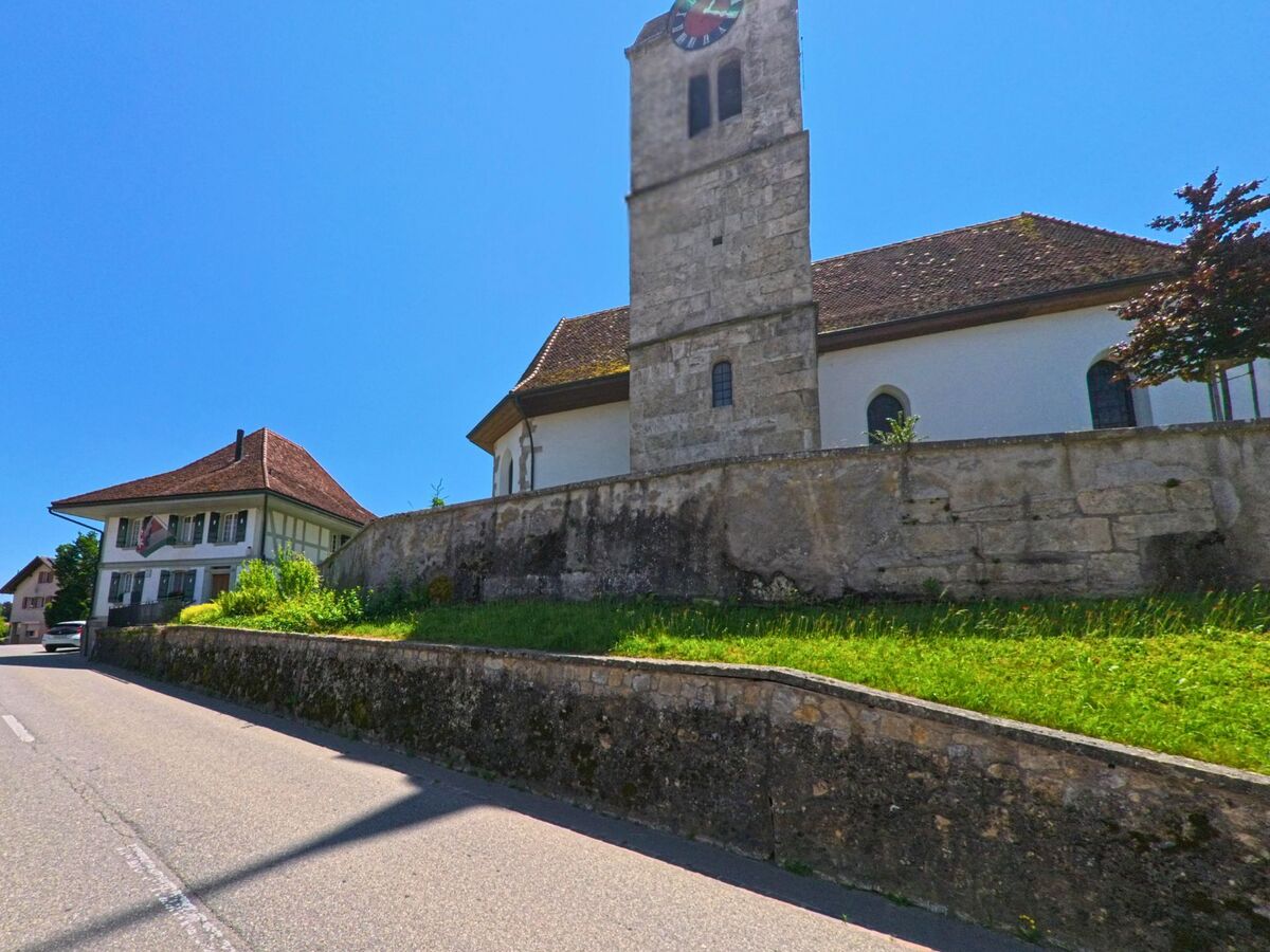 In Messen (3254, Kanton Solothurn) erhebt sich eine Kirche mit markantem Uhrturm und Umfassungsmauer über einer ansteigenden Strasse mit historischen Wohnhäusern.