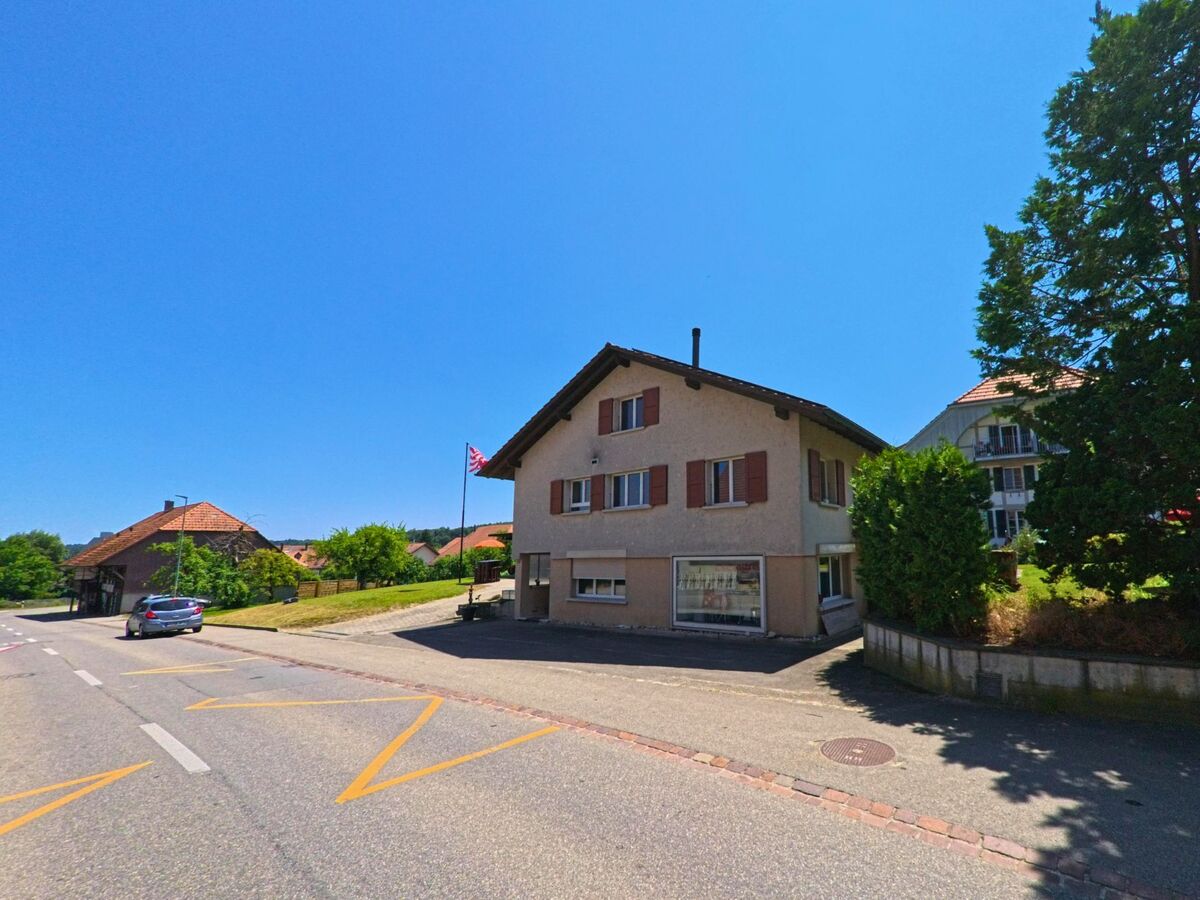 In Messen (3254, Kanton Solothurn) steht ein schlichtes Wohnhaus mit braunen Fensterläden und Ladeneingang an einer Durchgangsstrasse im Dorfzentrum.