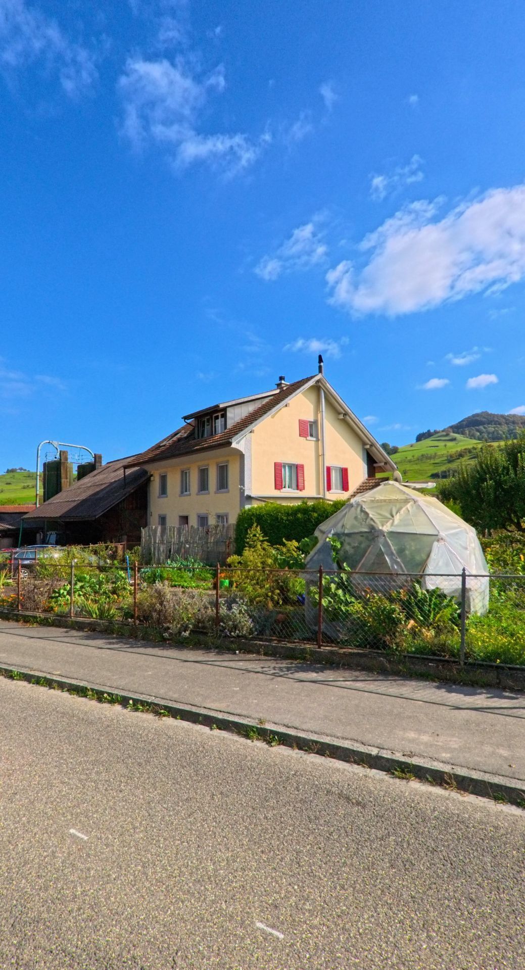 Ein Wohnhaus mit roten Fensterlaeden und einem kleinen Garten mit Gewaechshaus steht in Mettauertal (5273, Kanton Aargau) vor einer huegeligen Landschaft.