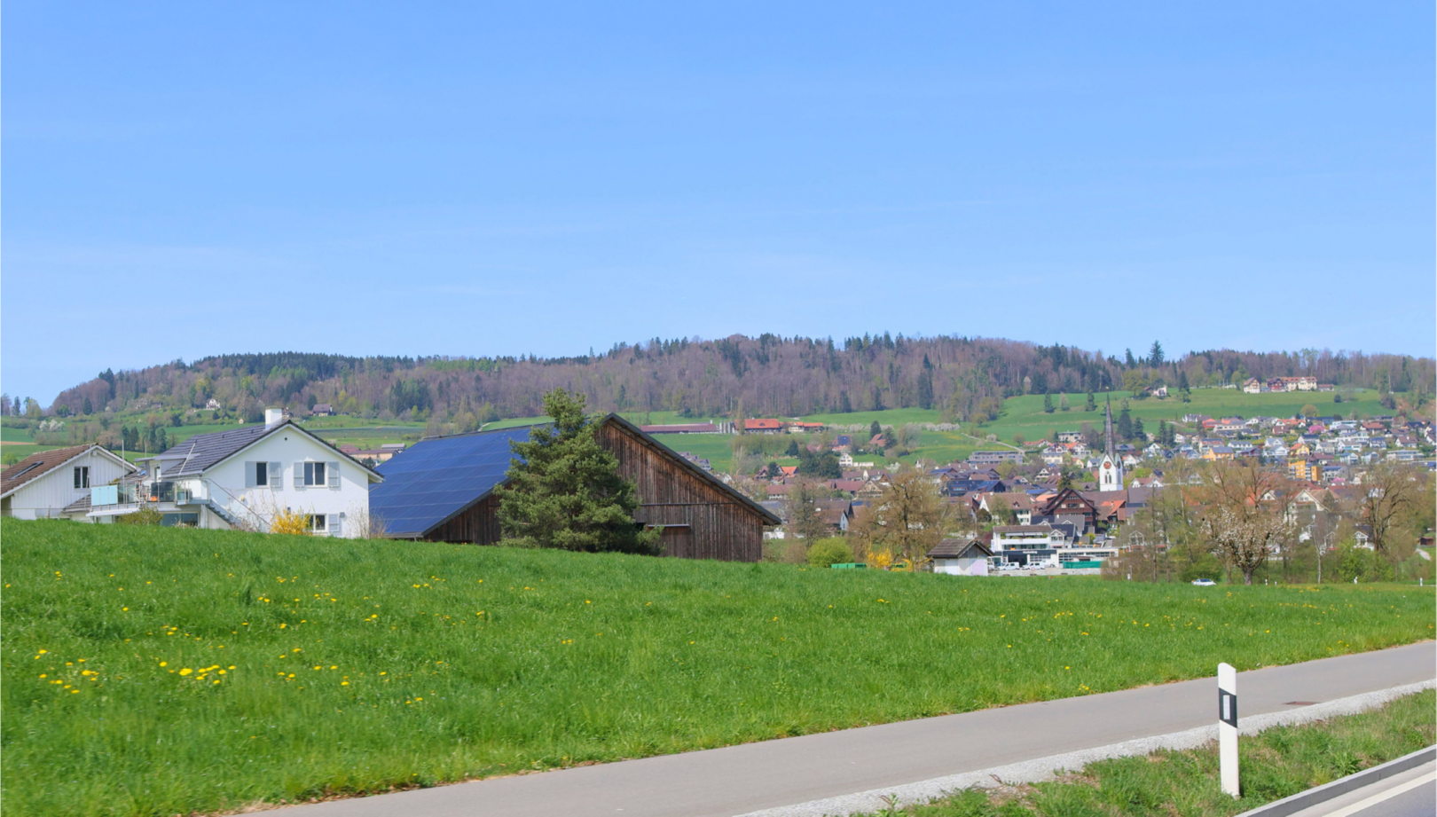 Am Ortsrand von Mettmenstetten, PLZ 8934 im Kanton Zürich, stehen ein Wohnhaus mit Solarpanels und eine Scheune vor der Kulisse des Dorfzentrums mit Kirche und Hügeln.