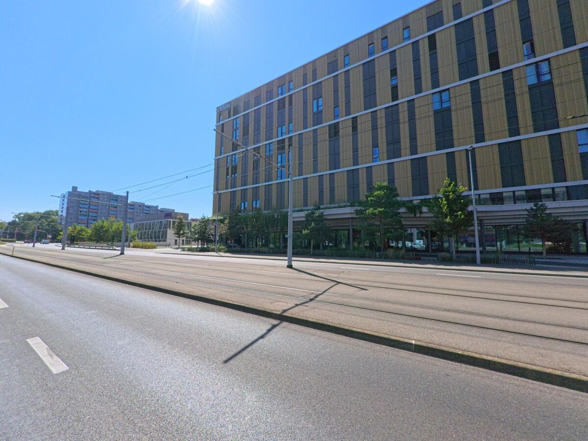 In Meyrin (1216, Kanton Genf) verlaeuft eine breite Strasse mit Tramgleisen entlang eines modernen Hochhauses mit Holzfassade.