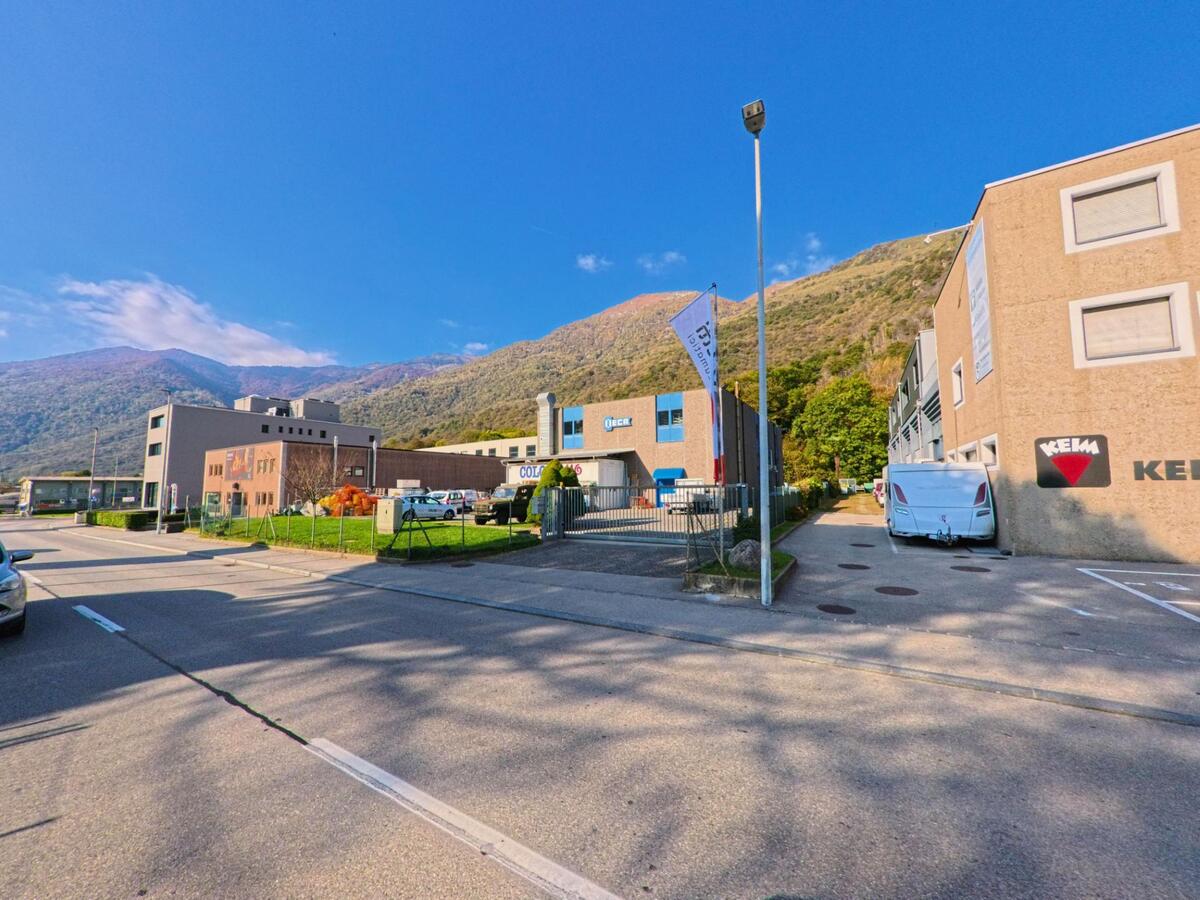 Zwischen mehreren Betriebsgebäuden und Zufahrten zeigt sich ein Gewerbeareal in Mezzovico-Vira (6805, Kanton Tessin), eingerahmt von bewaldeten Berghängen und klarem Himmel.