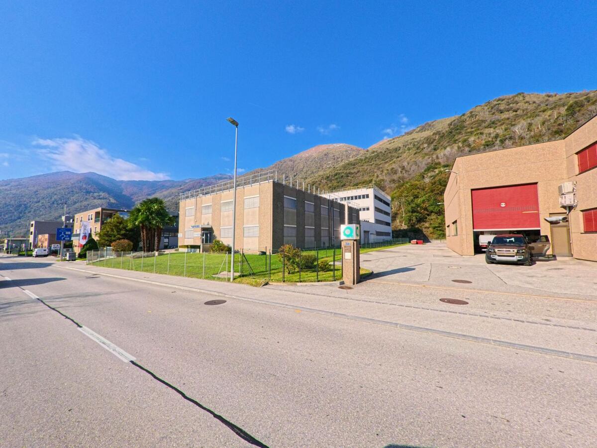 Entlang einer breiten Zufahrtsstrasse liegen funktionale Gewerbebauten in Mezzovico-Vira (6805, Kanton Tessin), eingebettet zwischen Grünflächen und steilen Berghängen unter klarem Himmel.
