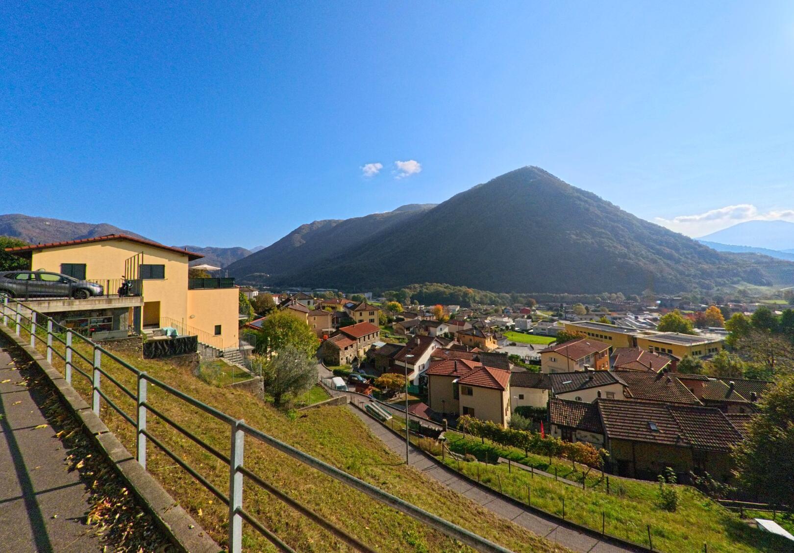 Von einem erhöhten Weg aus zeigt sich der Blick über Wohnhäuser und Gärten von Mezzovico-Vira (6805, Kanton Tessin) vor einem bewaldeten Berghang unter klarem Himmel.