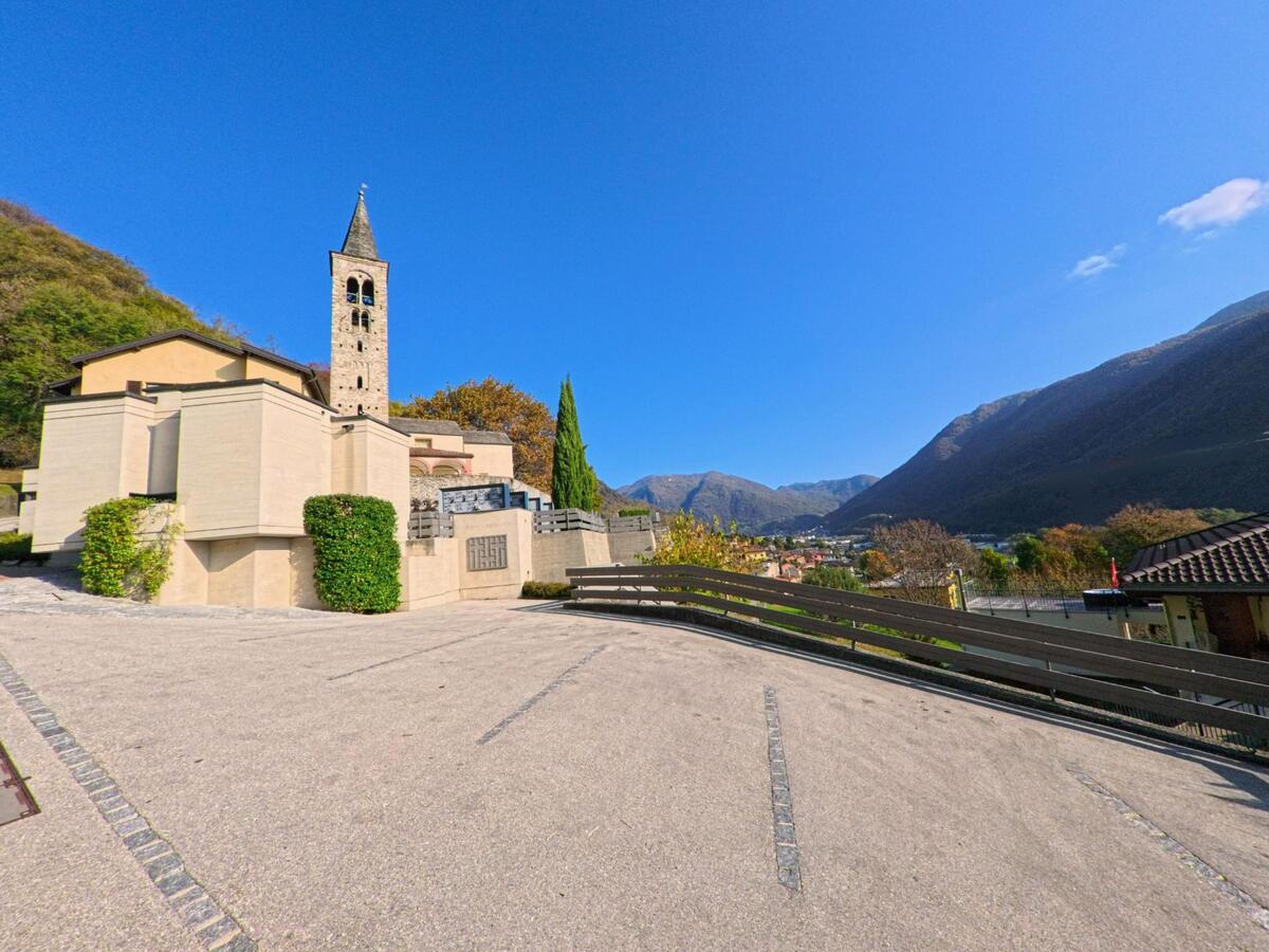 Neben einem markanten Kirchturm öffnet sich ein gepflasterter Platz mit Blick ins Tal von Mezzovico-Vira (6805, Kanton Tessin), eingerahmt von Gebäuden und bewaldeten Berghängen.