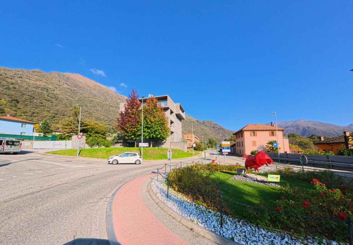 Am Rand eines Kreisels mit bepflanzter Mittelinsel zeigt sich eine Verkehrssituation in Mezzovico-Vira (6805, Kanton Tessin), eingerahmt von Wohnhäusern, Gewerbebauten und bewaldeten Berghängen.
